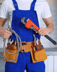 Plombier en salopette bleue tenant une clé à molette rouge et un flexible de douche. Ceinture à outils en cuir avec outils.