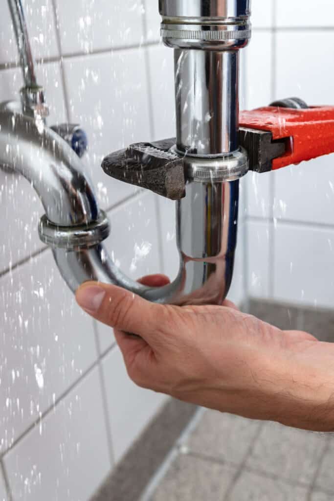 Main d'homme serrant un siphon chromé avec une clé à molette rouge pour réparer une fuite d'eau éclaboussant sur des carreaux blancs.